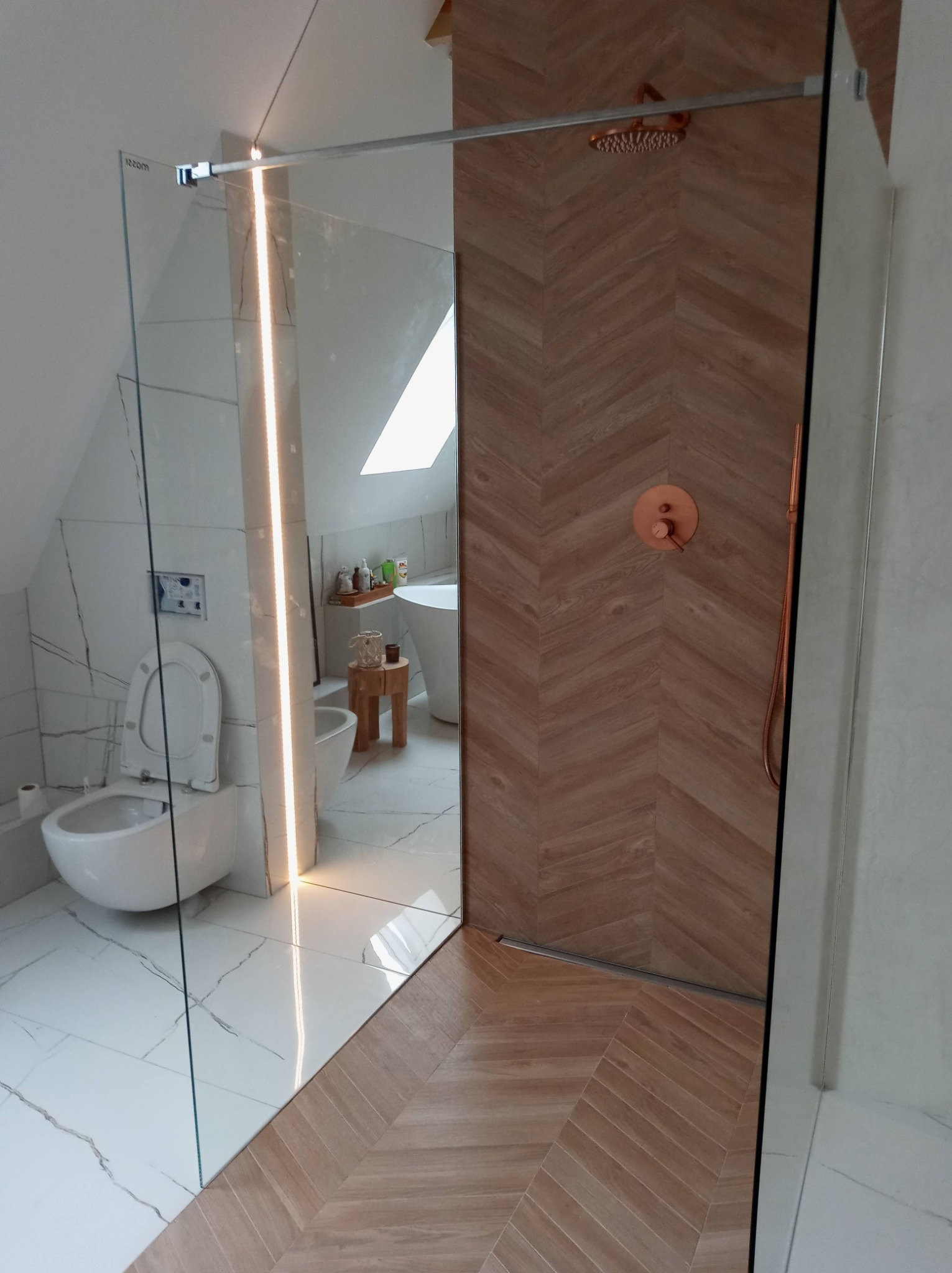 Nowoczesna łazienka z kabiną prysznicową typu walk-in, wykończona płytkami imitującymi drewno ułożonymi w jodełkę, z armaturą w kolorze miedzianym i widocznym fragmentem toalety z podtynkową spłuczką.