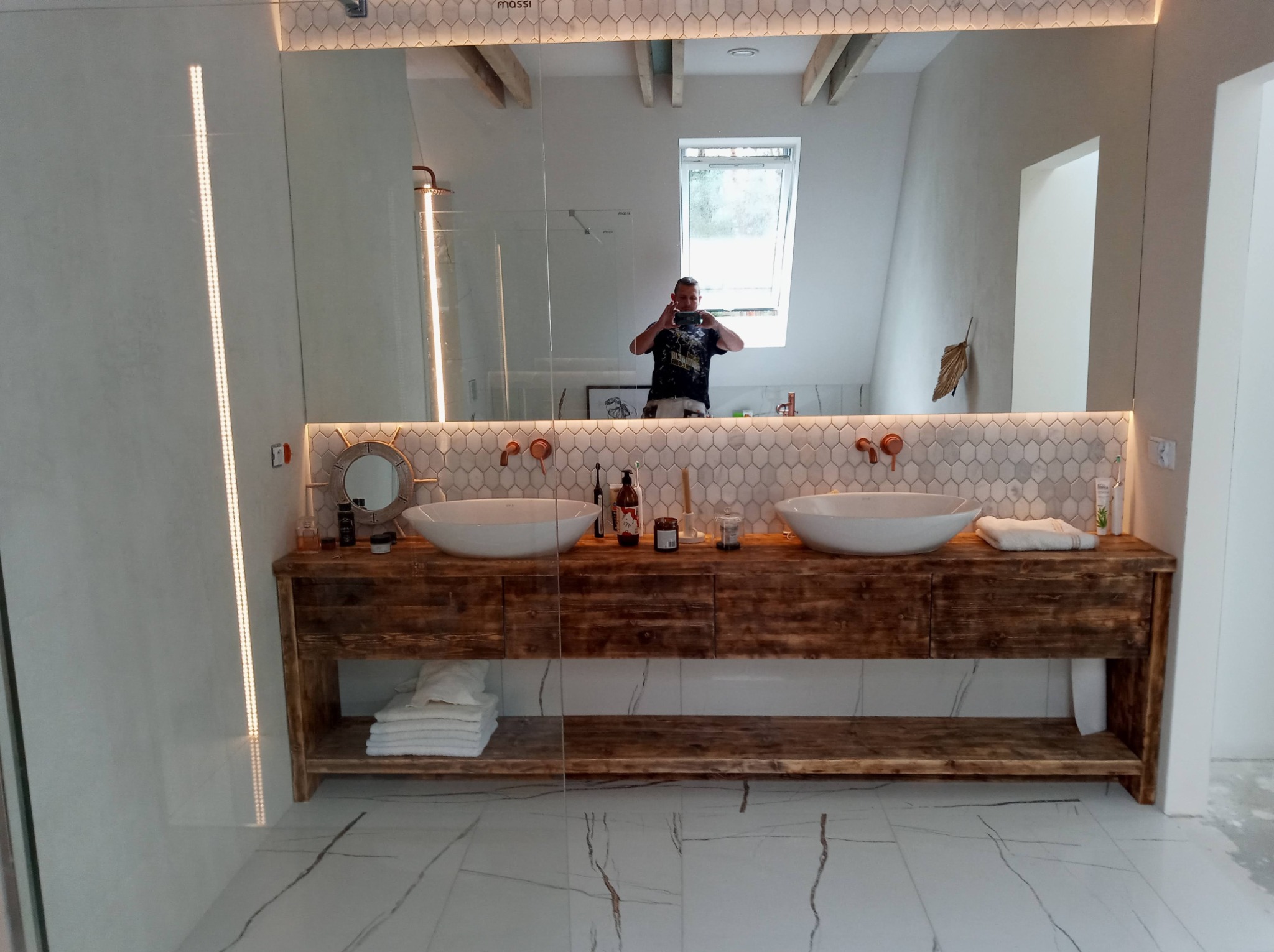 Nowoczesna łazienka z drewnianą szafką pod umywalki nablatowe, białymi płytkami heksagonalnymi na ścianie i marmurową podłogą, odbicie osoby robiącej zdjęcie w lustrze.