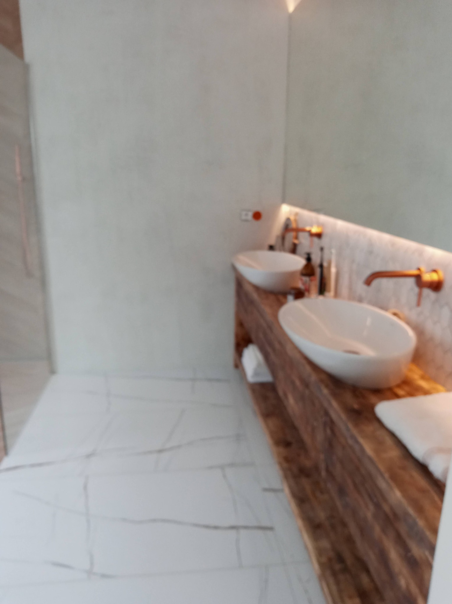 Nowoczesna łazienka z dwoma umywalkami nablatowymi o owalnym kształcie, osadzonymi na drewnianym blacie o wyraźnej strukturze, z miedzianymi bateriami i podświetlanym lustrem, białe płytki...