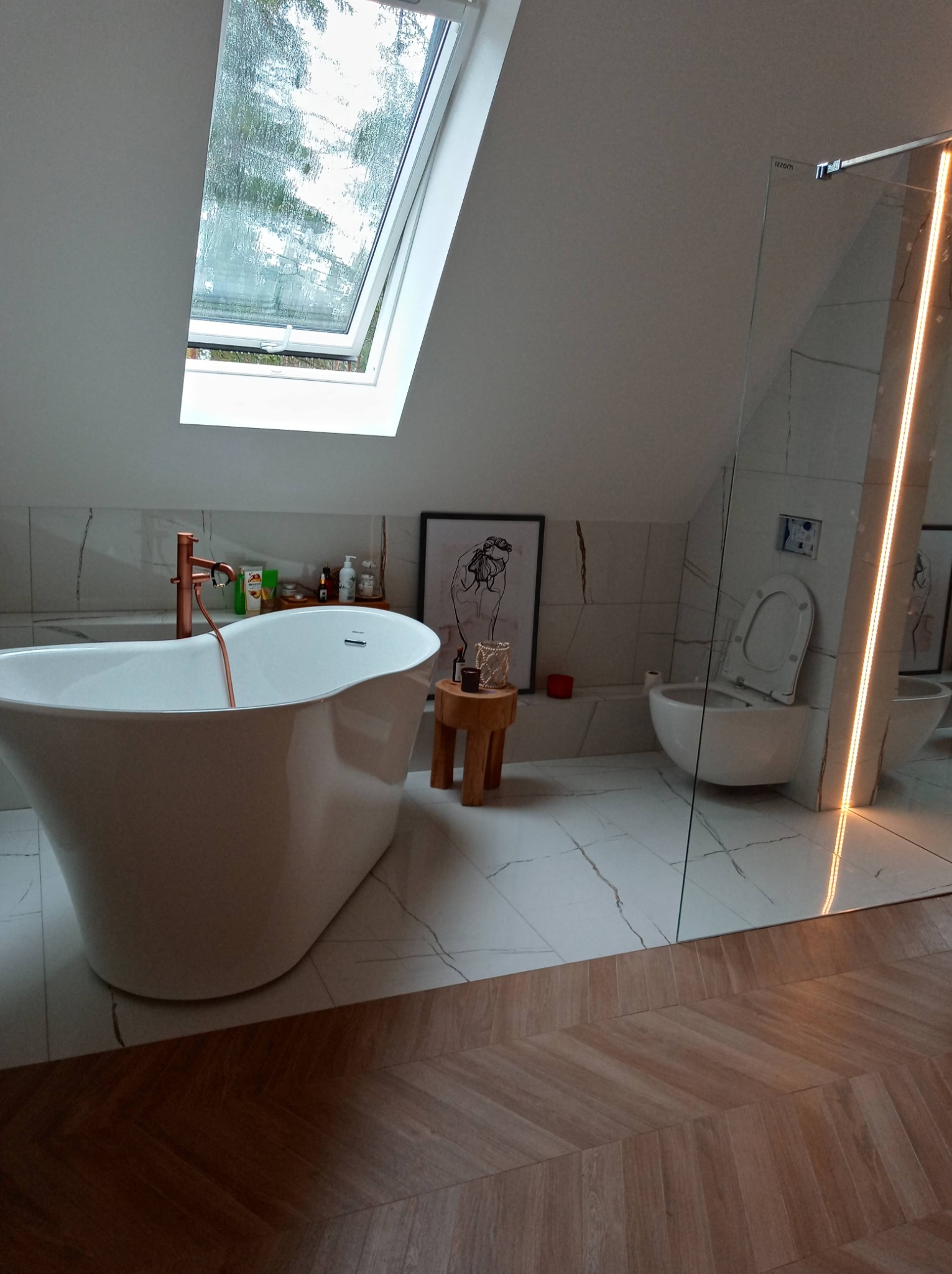 Nowoczesna łazienka na poddaszu z wolnostojącą wanną, baterią w kolorze miedzi, toaletą podwieszaną i kabiną prysznicową ze szklaną ścianą z podświetleniem LED.