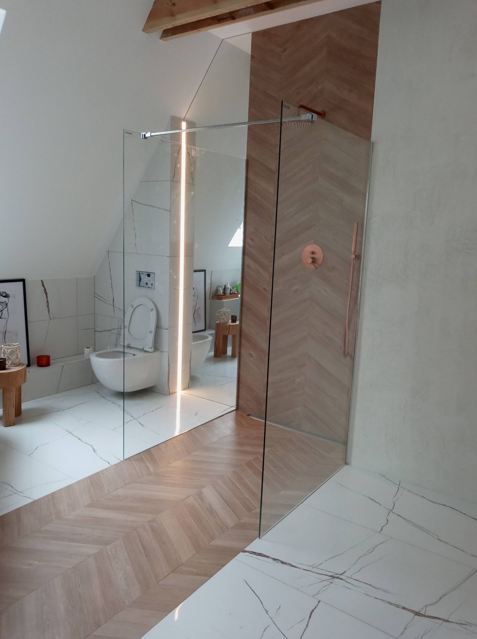 Nowoczesna łazienka na poddaszu z kabiną prysznicową typu walk-in, wykończona płytkami imitującymi marmur i drewnem ułożonym w jodełkę, widoczne elementy armatury w kolorze miedzianym.