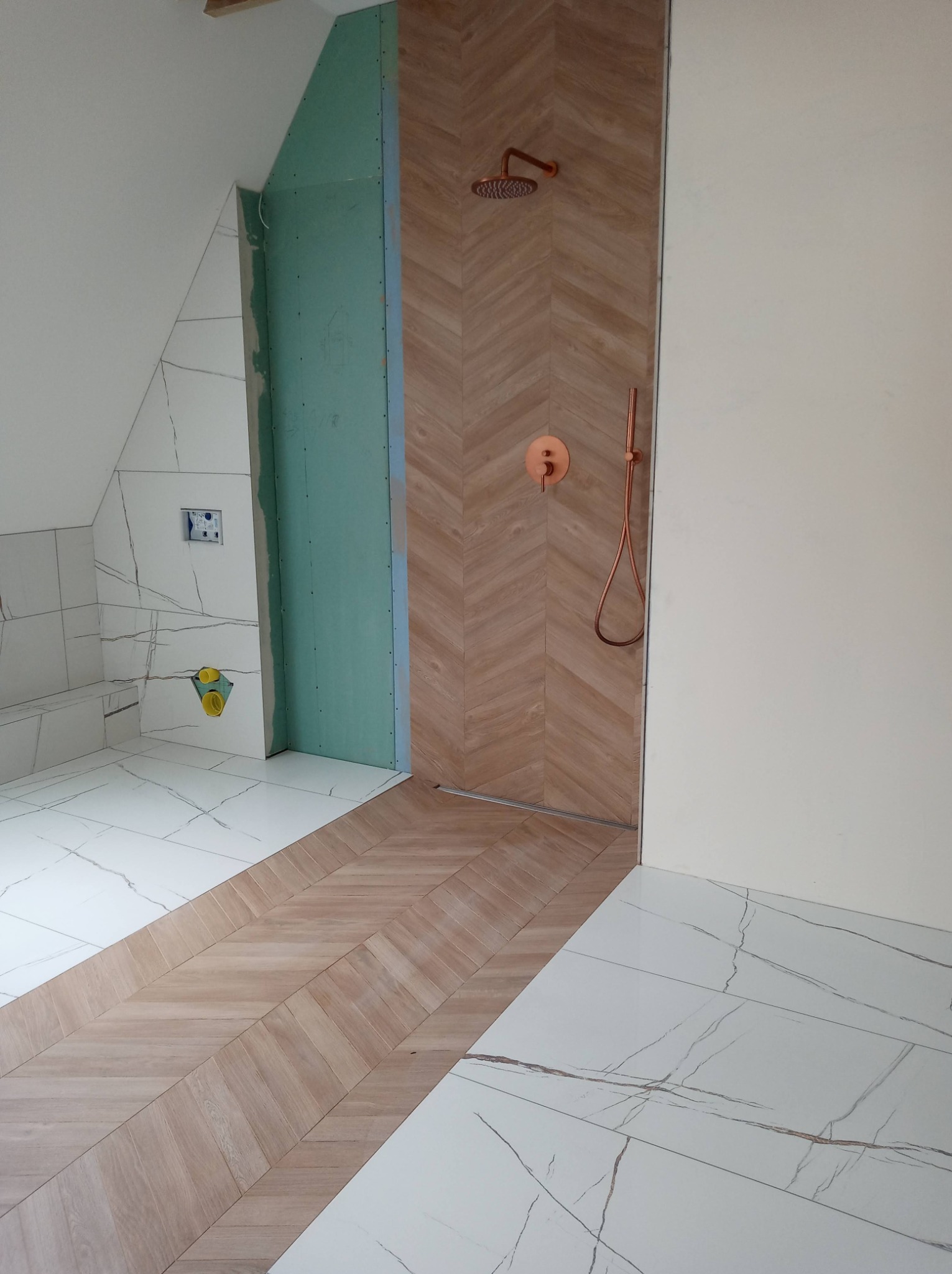 Wnętrze łazienki w trakcie remontu, widoczne białe płytki imitujące marmur na podłodze i ścianach, fragment ściany z drewnopodobnymi płytkami ułożonymi w jodełkę, miedziana armatura prysznicowa...