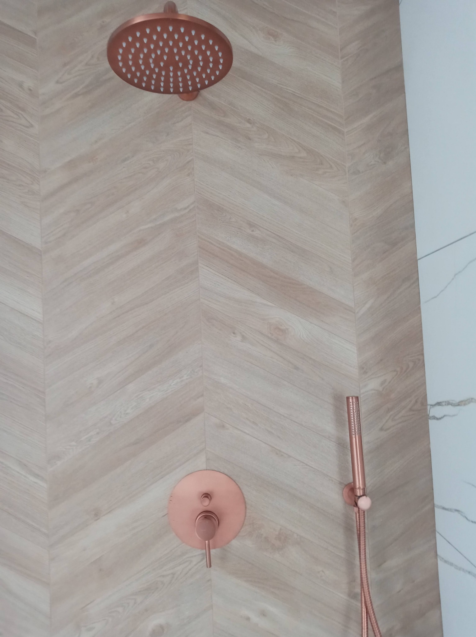 Nowoczesna łazienka z miedzianą armaturą: deszczownica, bateria i słuchawka prysznicowa na ścianie wyłożonej płytkami imitującymi drewno ułożonymi w jodełkę, obok fragment ściany z białego marmuru.