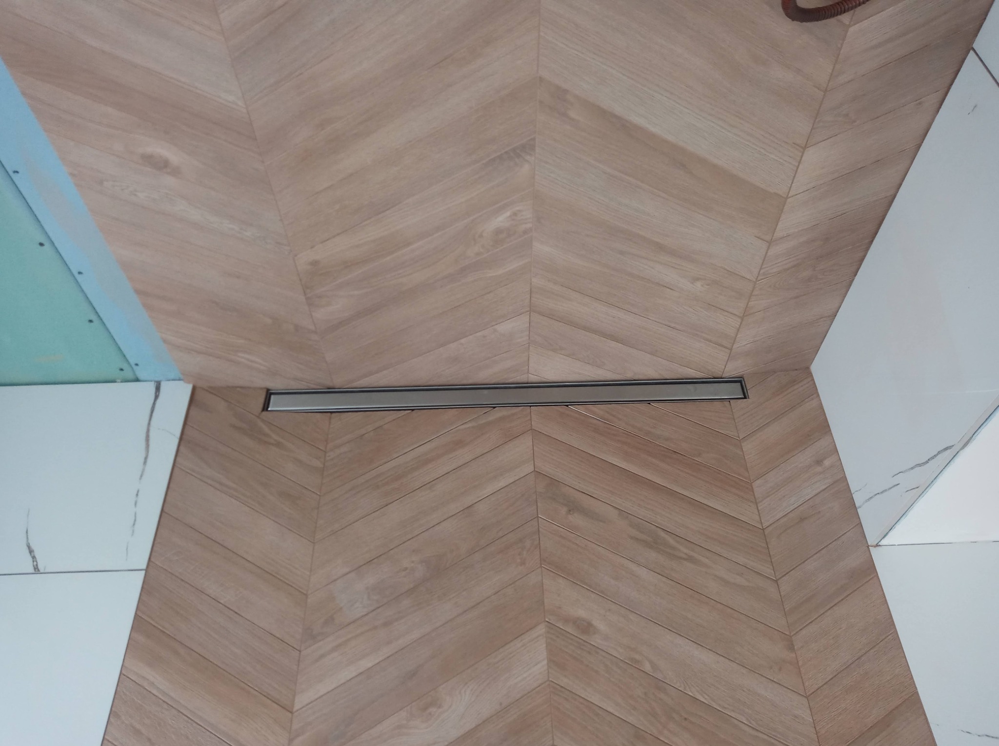 Ułożone w jodełkę płytki podłogowe imitujące drewno w łazience, z odpływem liniowym i fragmentem ściany z płyt gipsowo-kartonowych oraz białych płytek imitujących marmur.