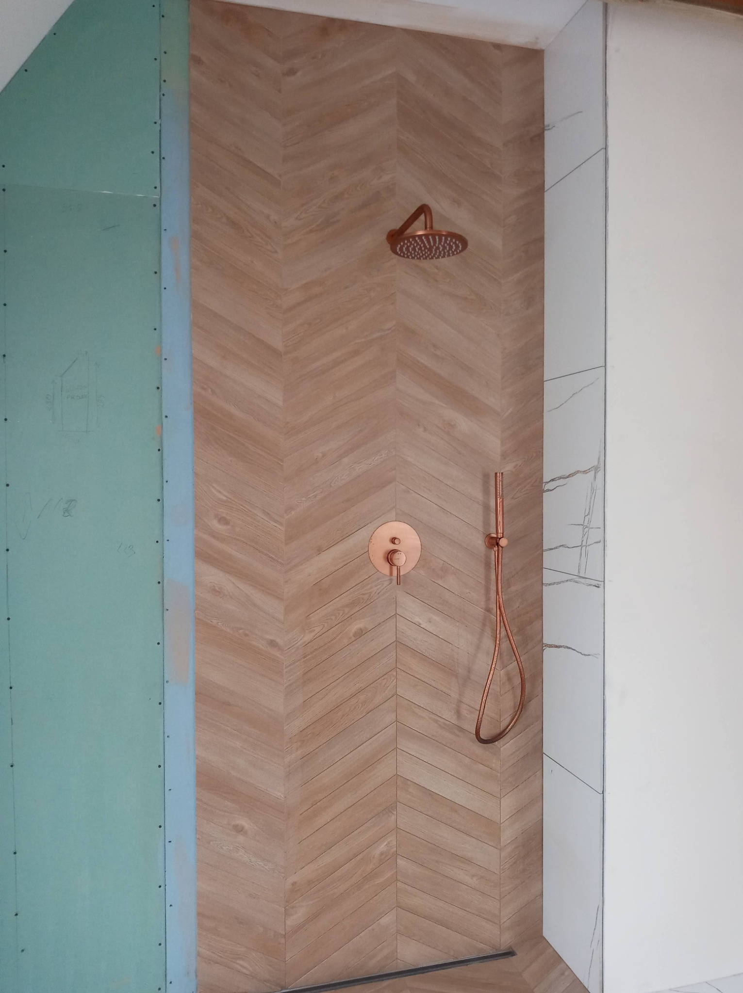 Niedokończona łazienka z prysznicem: ściana wyłożona płytkami imitującymi drewno w jodełkę, miedziana armatura, płyta gipsowo-kartonowa na ścianie obok.