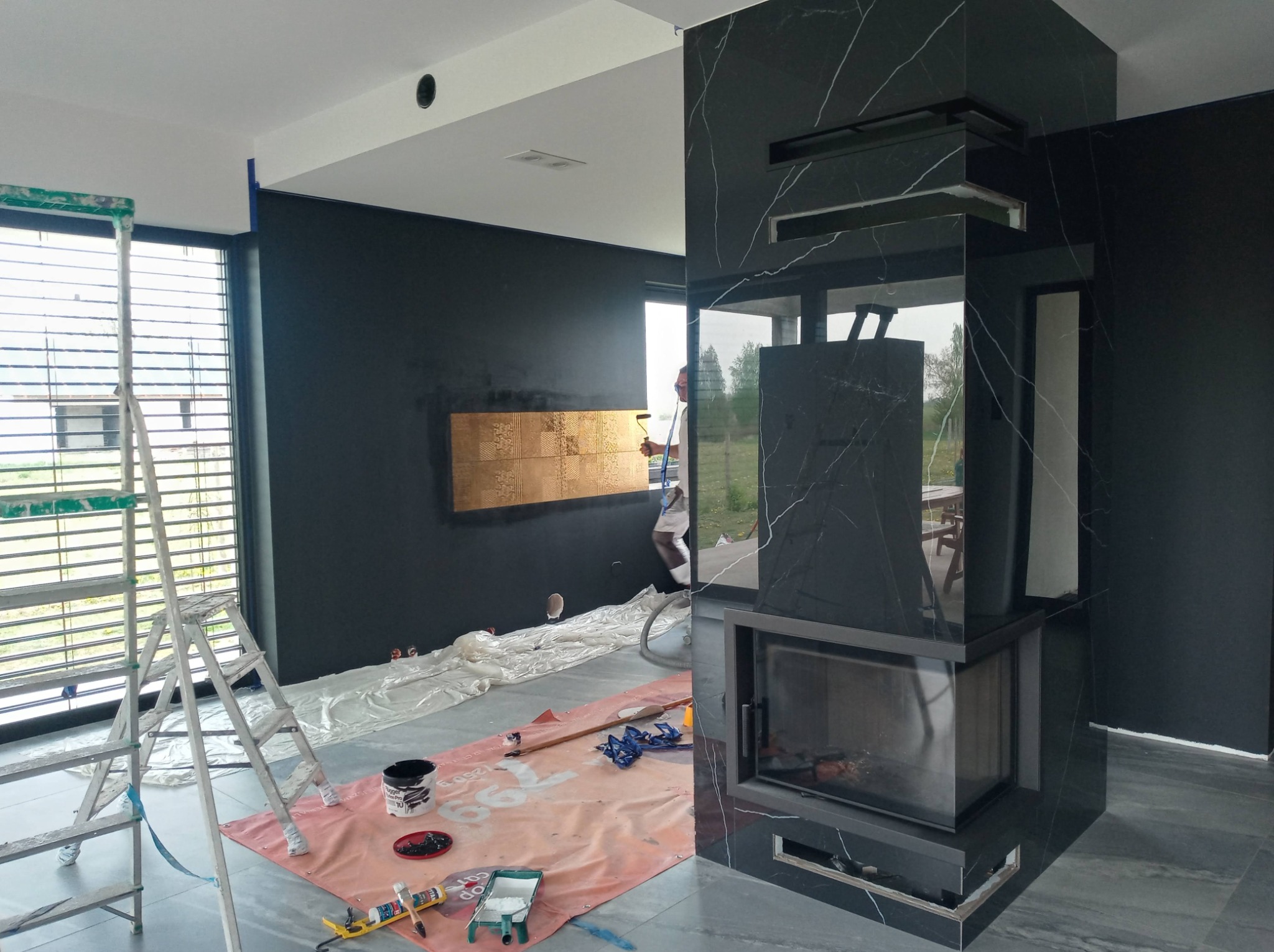 Wnętrze w trakcie remontu z czarnymi ścianami, marmurowym kominkiem i rozłożonymi foliami ochronnymi na podłodze. Malarz w tle maluje ścianę na złoty kolor.