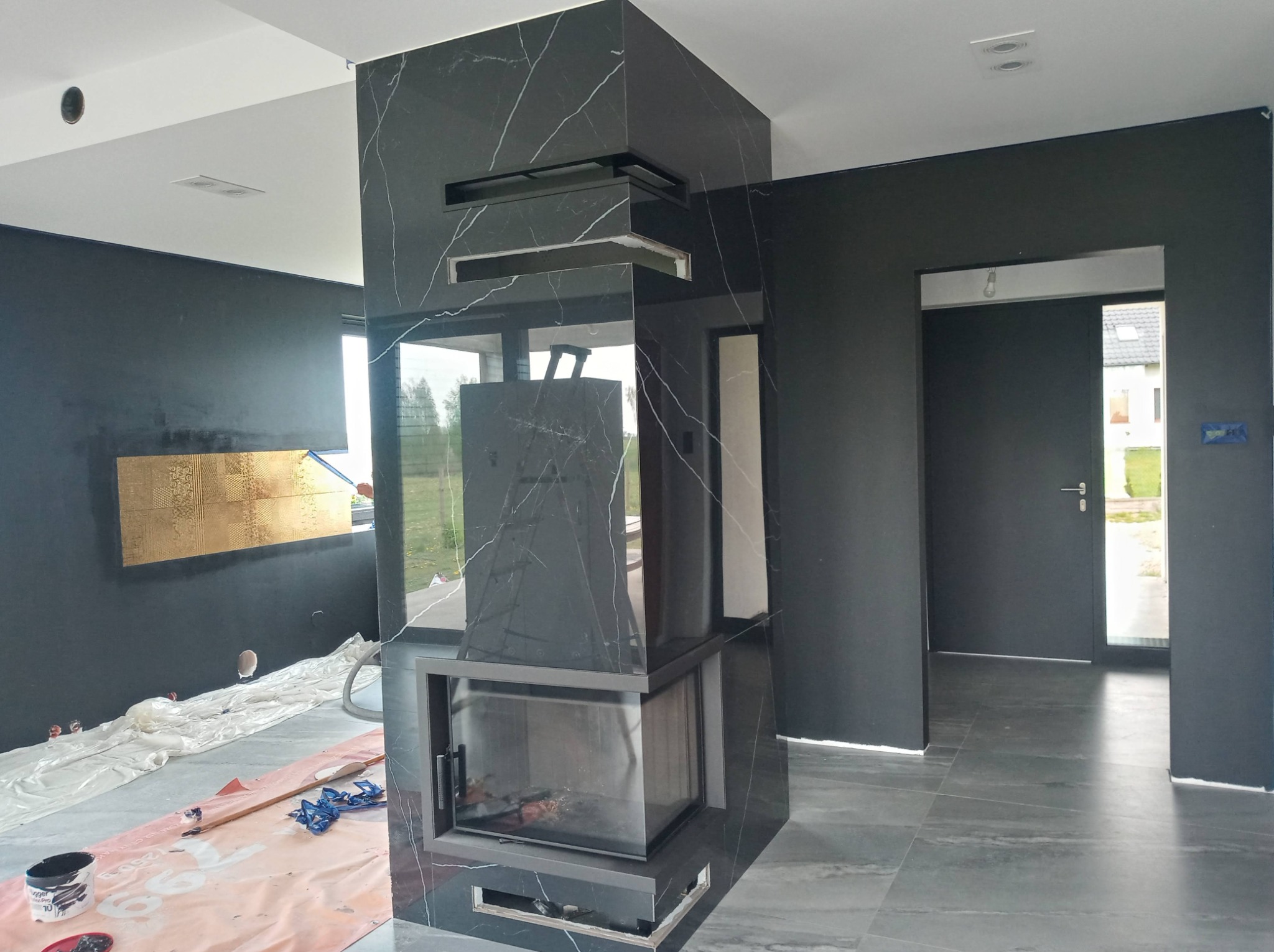 Wnętrze w trakcie wykańczania z czarnym kominkiem obłożonym płytami imitującymi marmur, zabezpieczona podłoga, malowane na czarno ściany i widoczny fragment ozdobnej ściany w kolorze złota.