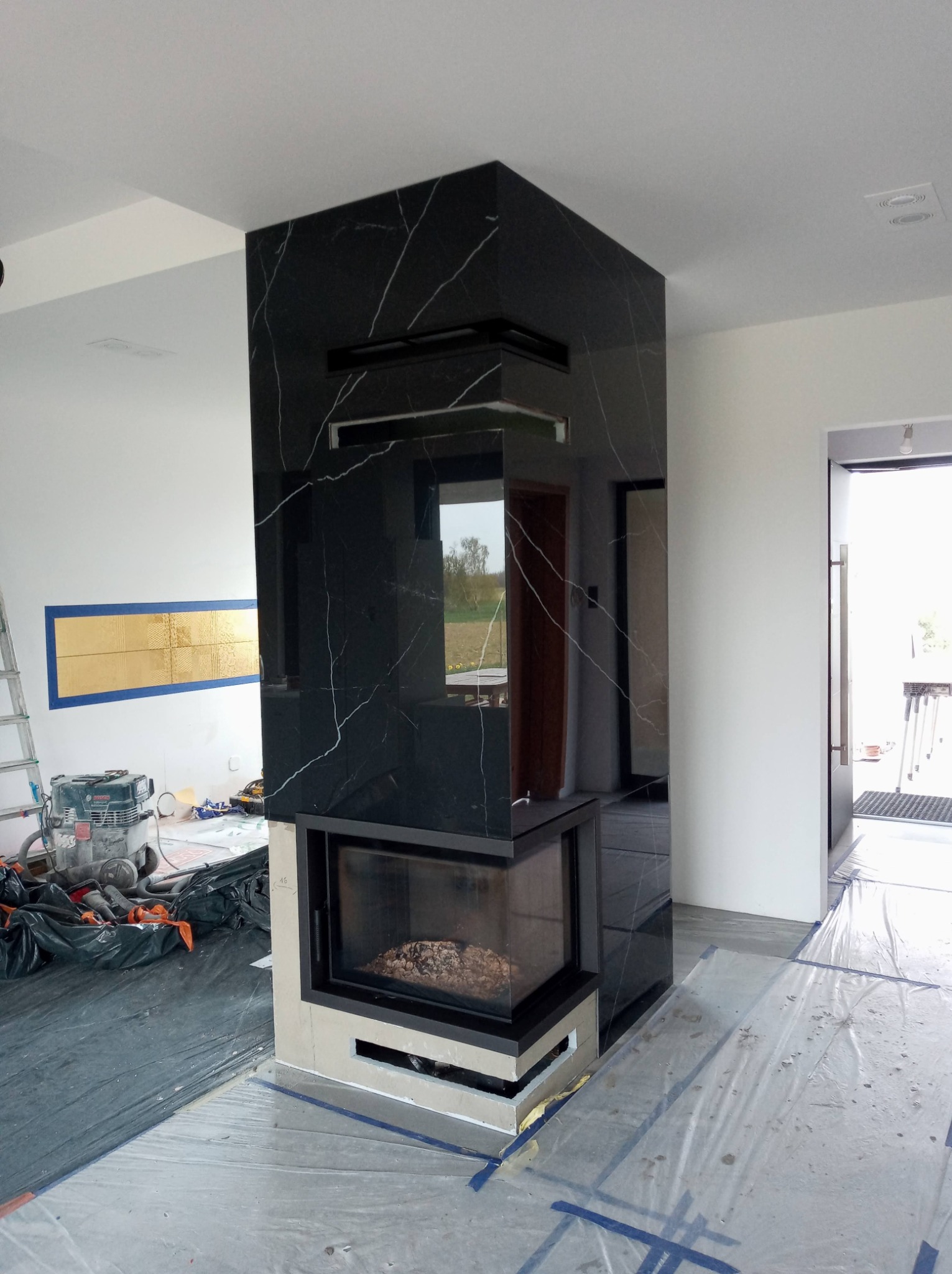 Wnętrze w trakcie prac wykończeniowych, widoczny nowoczesny kominek obłożony czarnym marmurem z białymi żyłami, zabezpieczona folią podłoga, drabina i odkurzacz przemysłowy w tle.