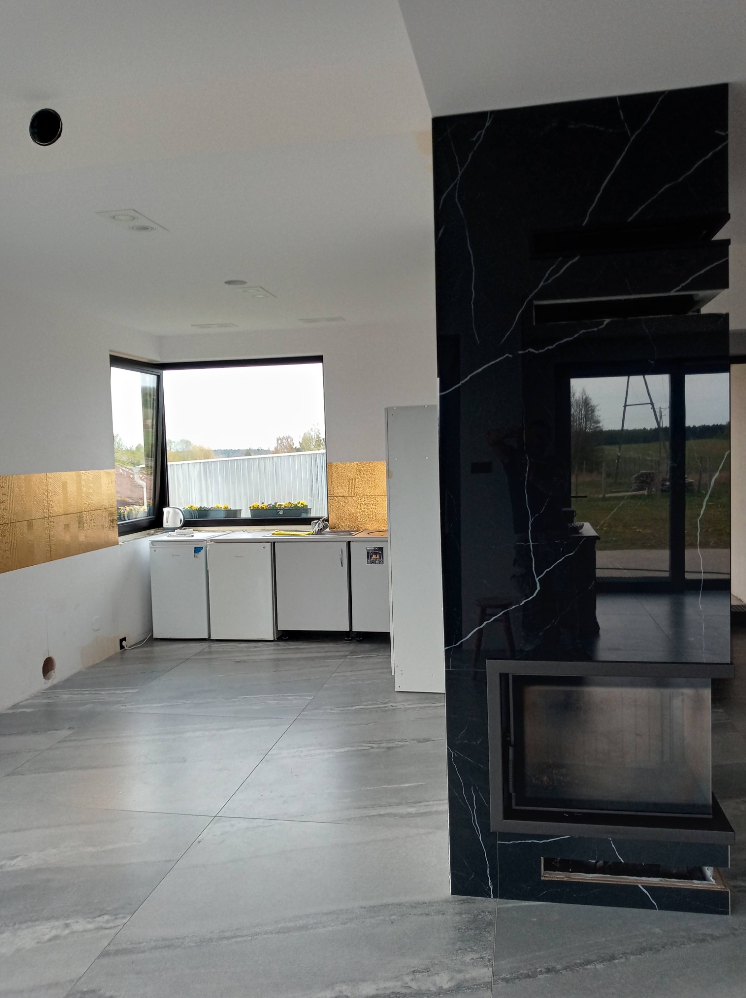 Wnętrze w trakcie wykańczania: szare płytki podłogowe, czarny kominek z marmurowym wykończeniem, fragment ściany z złotymi płytkami, okno z widokiem na zewnątrz, białe lodówki pod oknem.
