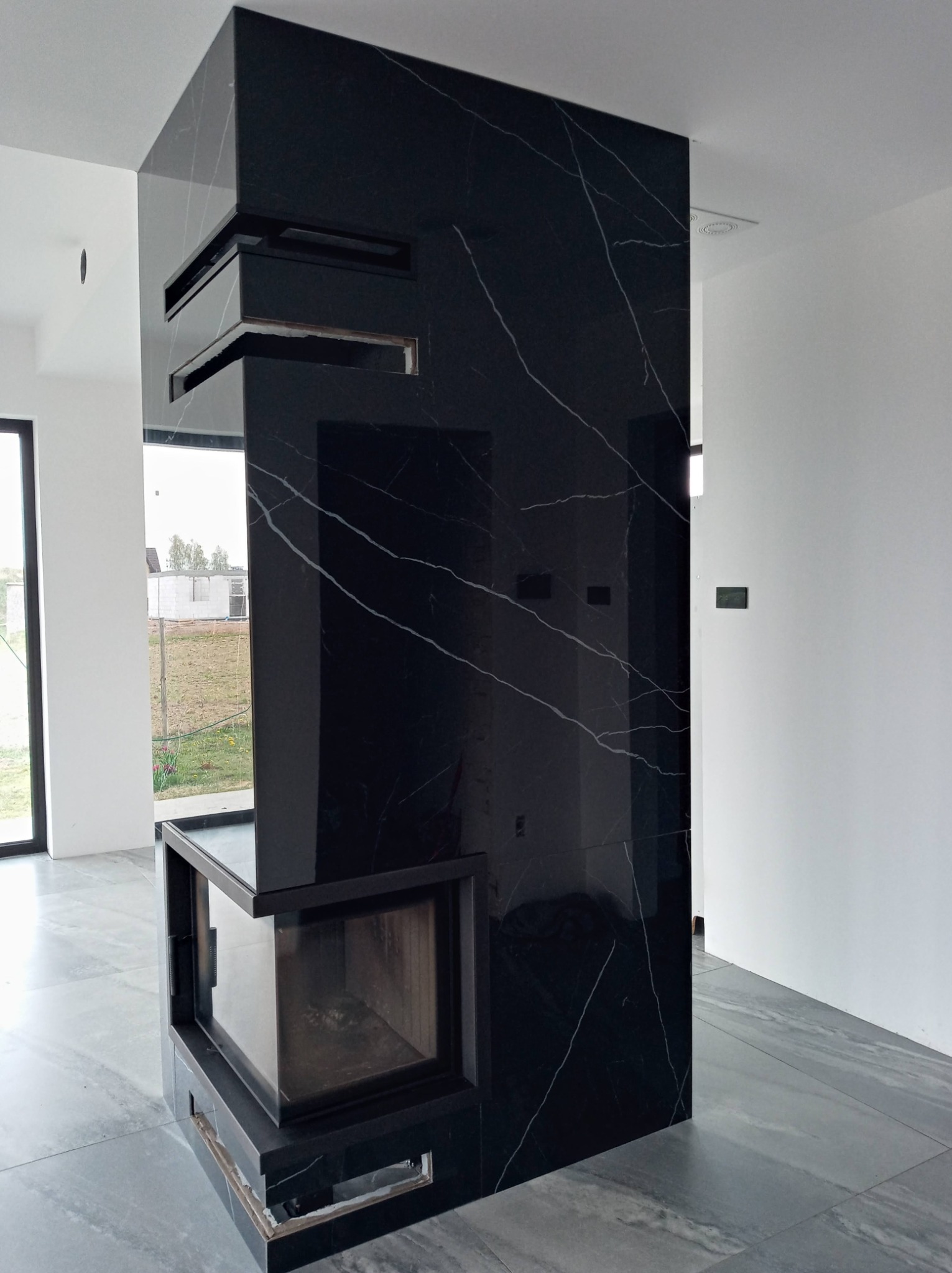 Nowoczesny kominek obłożony czarnym marmurem z białymi żyłami, widoczny w surowym wnętrzu z szarą podłogą i białymi ścianami, z widokiem na budowę za oknem.