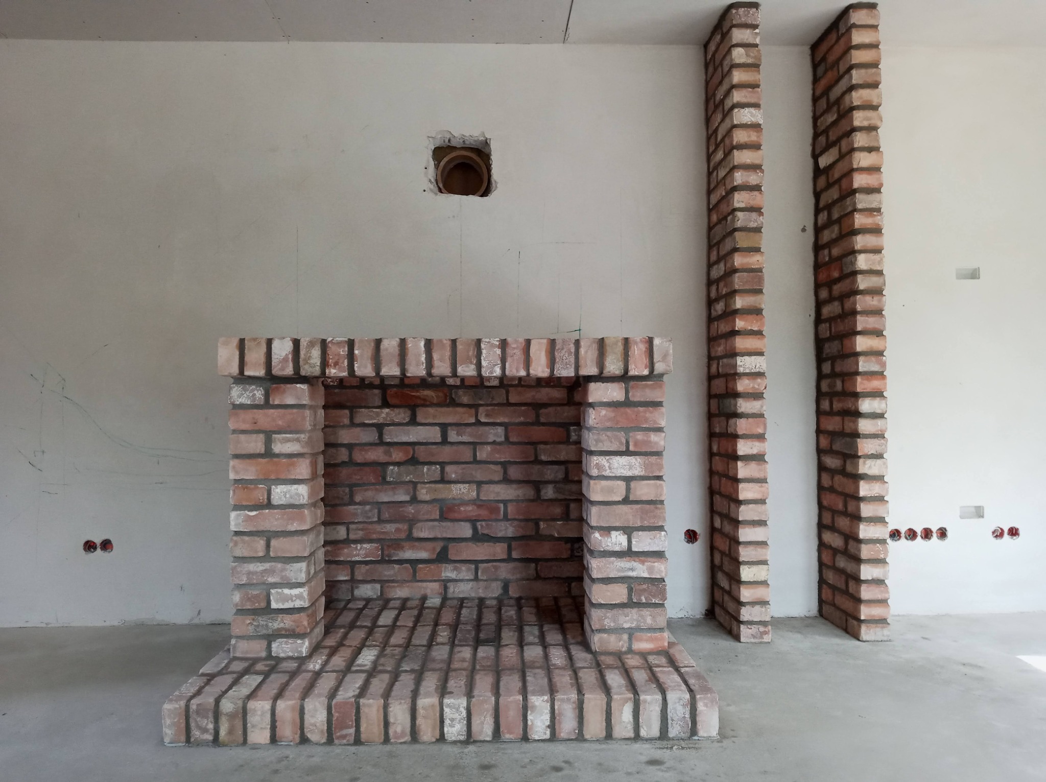 Wnętrze w trakcie prac wykończeniowych z częściowo wymurowanym kominkiem z cegły klinkierowej i fragmentami obudowy, widoczne niedokończone ściany i instalacje elektryczne.