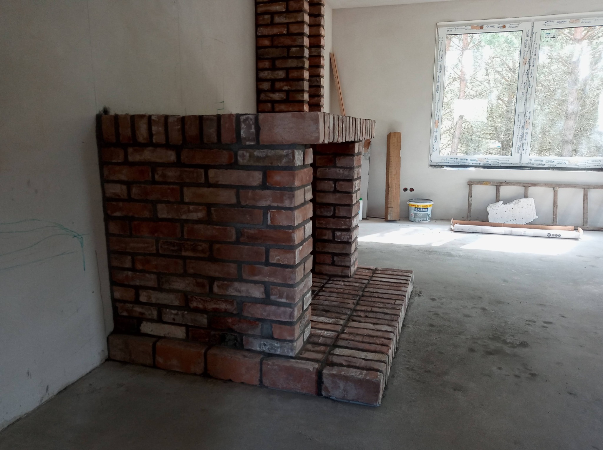 Wnętrze w trakcie prac wykończeniowych: murowany z czerwonej cegły kominek w surowym stanie, widoczne ślady zaprawy, na podłodze betonowa wylewka, w tle okno z widokiem na drzewa.