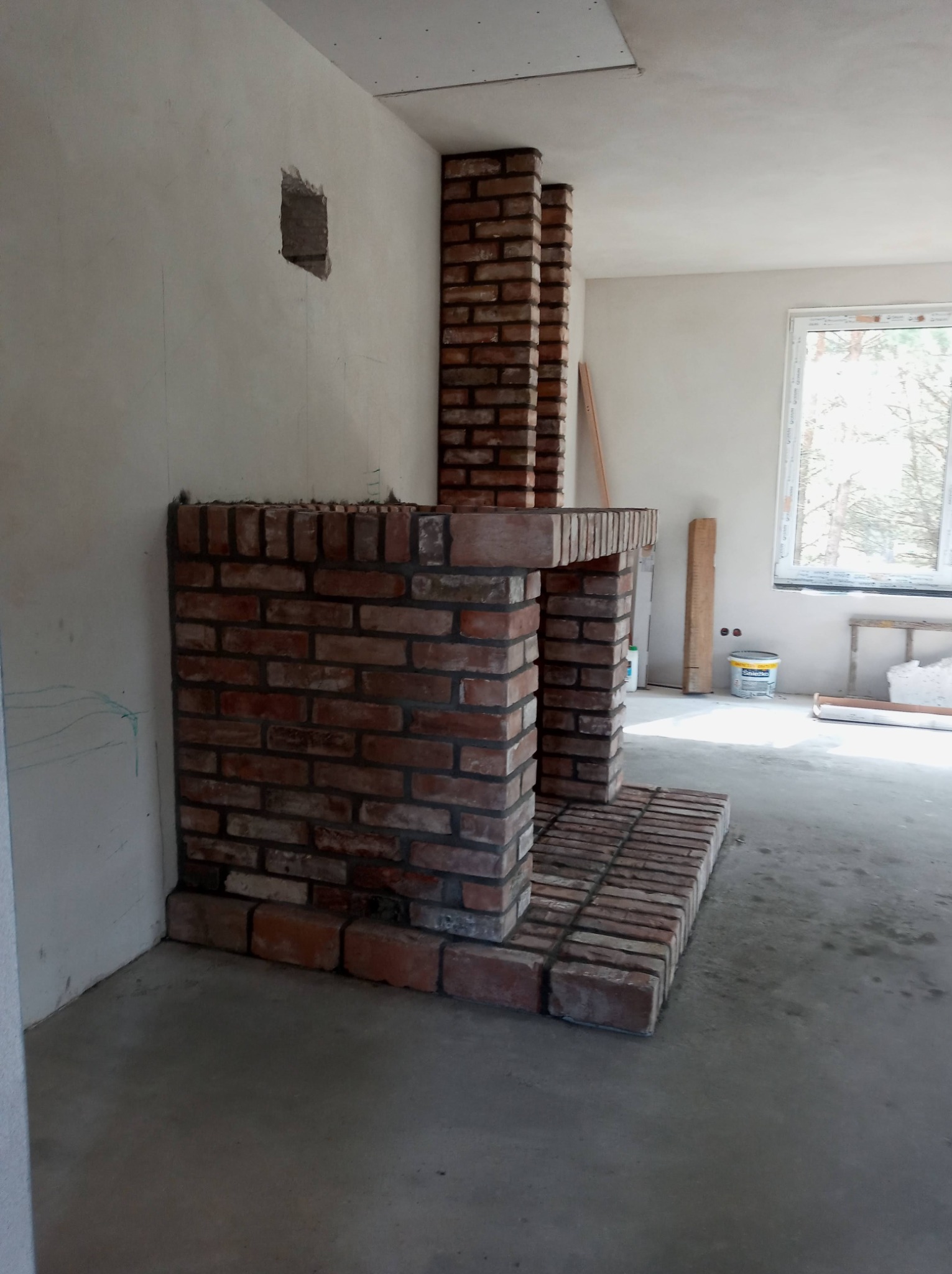 Murowany kominek z cegły w surowym stanie, widoczne niedoskonałości spoin, ściany w trakcie wykończenia, otwór w ścianie, okno z widokiem na drzewa w tle.