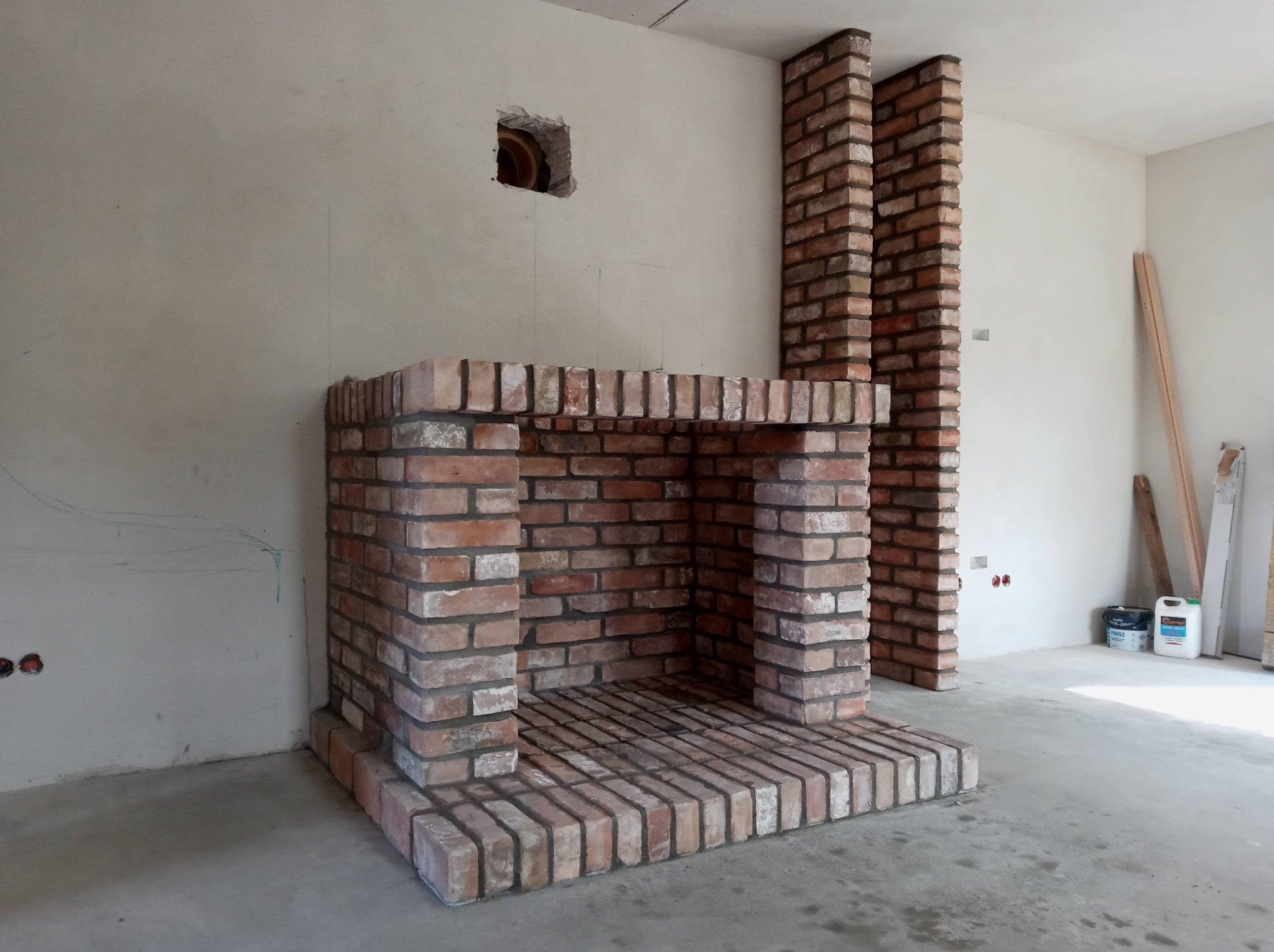 Murowany kominek z cegły klinkierowej w surowym wnętrzu, widoczny otwór wentylacyjny w ścianie i przygotowane instalacje elektryczne, etap budowy.
