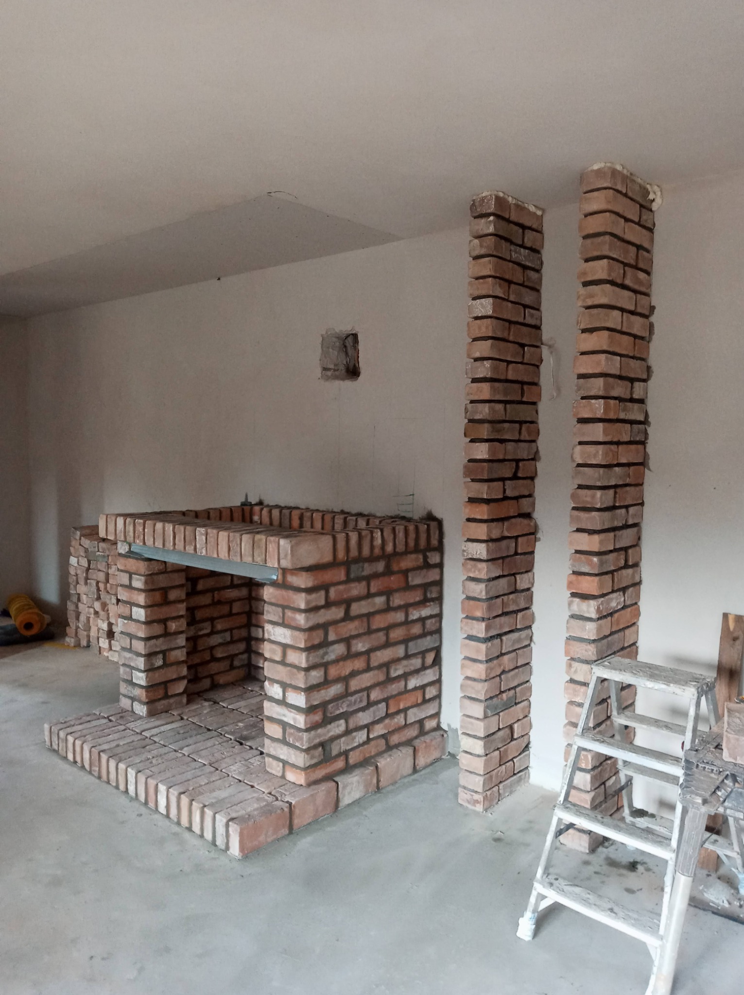 Wnętrze w trakcie budowy: murowany kominek z czerwonej cegły, wsparty na metalowej belce, z dwoma kolumnami z cegły po bokach, drabina i narzędzia w tle, surowy stan wykończenia.