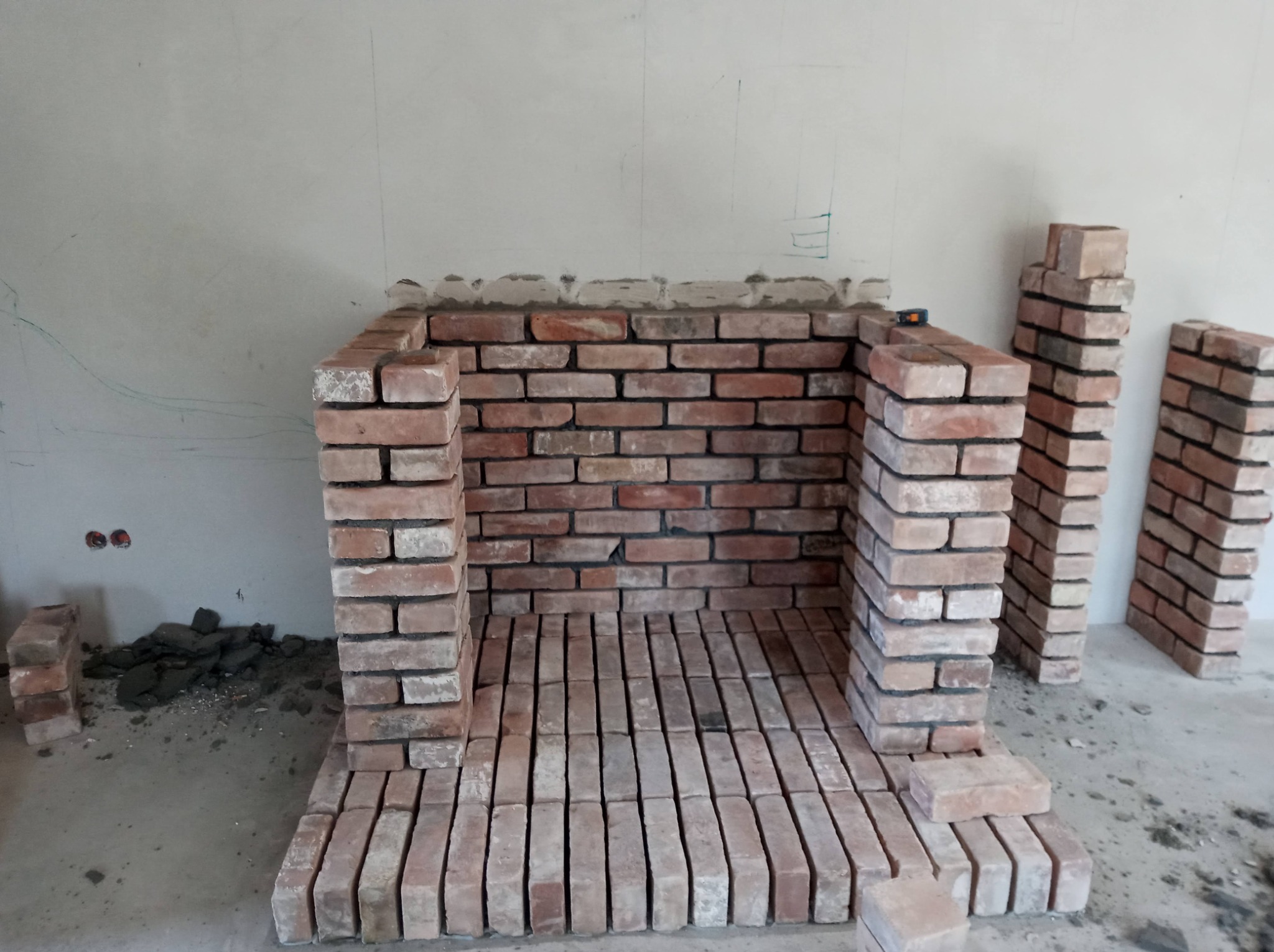 Wnętrze kominka w trakcie budowy z czerwonej cegły, widoczne niedociągnięcia w spoinach i resztki zaprawy na podłodze, ściana z zaznaczonymi liniami pomocniczymi.