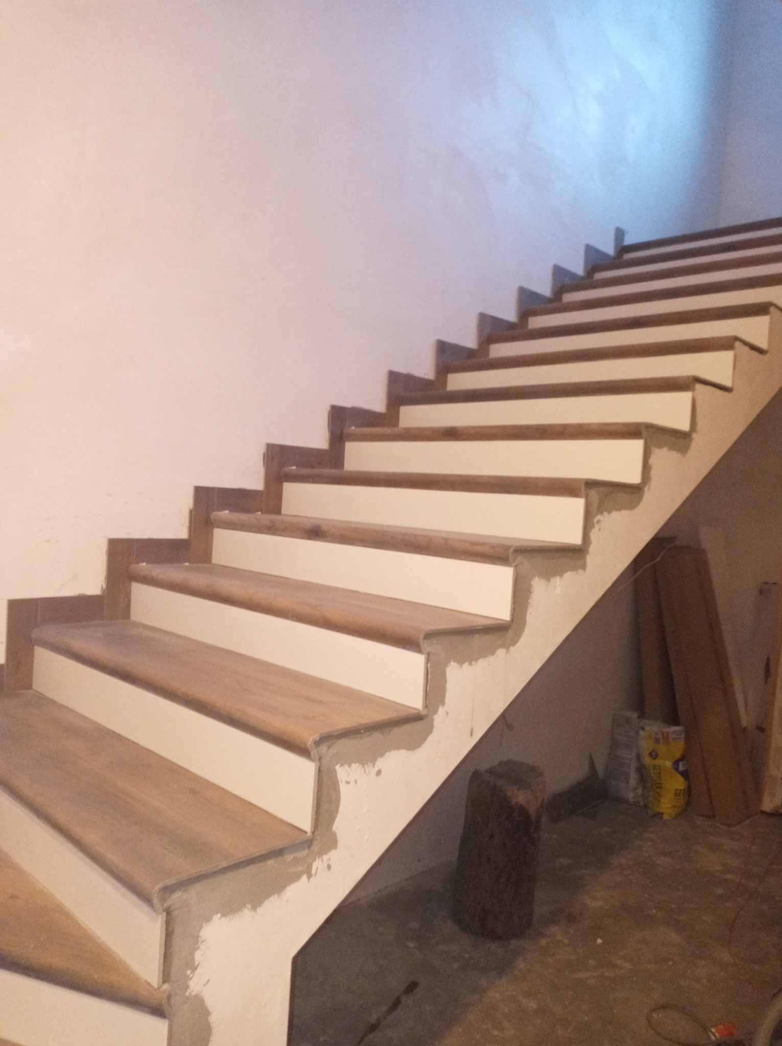 Drewniane schody z białymi podstopnicami w trakcie wykończenia, widoczne ślady zaprawy i niedomalowane fragmenty, w tle worki i deski.