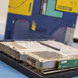 INsobi Konrad Sobiecki - Otwarta obudowa zakurzonej konsoli Microsoft XBOX One S, widoczne elementy wewnętrzne przed czyszczeniem, w tle pudełko konsoli w edycji specjalnej.