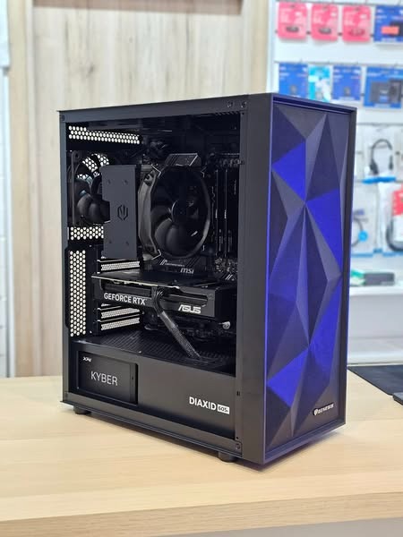 Komputer stacjonarny do gier w czarnej obudowie z niebieskimi akcentami, widoczna karta graficzna GeForce RTX i chłodzenie procesora, umieszczony na drewnianym biurku na tle półek sklepowych.