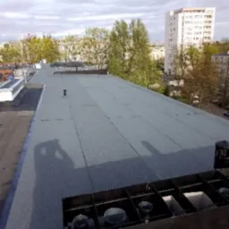 Widok z g&oacute;ry na płaski dach pokryty szarą papą termozgrzewalną, z widocznym cieniem osoby robiącej zdjęcie i elementami instalacji wentylacyjnej w prawym dolnym rogu, w tle budynki mieszkalne...