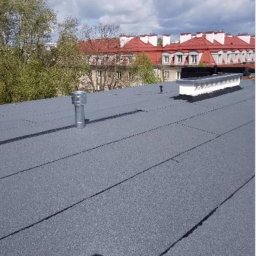 Miller. Bud. Oleh Miller - Szary, płaski dach pokryty papą termozgrzewalną z widocznymi kominkami wentylacyjnymi i świetlikiem dachowym na tle kamienicy z czerwoną dachówką i błękitnego nieba.