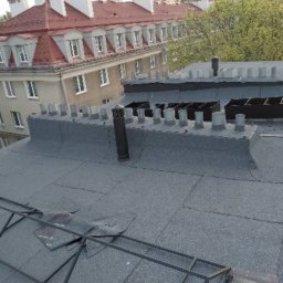 Miller. Bud. Oleh Miller - Widok z góry na płaski dach pokryty papą, z elementami wentylacyjnymi i kominem, w tle budynek mieszkalny z czerwonym dachem i zielone drzewa.