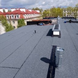 Miller. Bud. Oleh Miller - Wykończony, płaski dach pokryty papą termozgrzewalną z widocznymi kominkami wentylacyjnymi i elementami świetlików dachowych, w tle budynek mieszkalny z czerwonym dachem i błękitne niebo z chmurami.