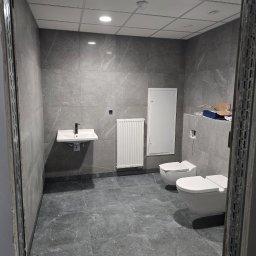 Miller. Bud. Oleh Miller - Szara łazienka z dwiema toaletami, umywalką i grzejnikiem, widok przez metalową futrynę w trakcie remontu, widoczne płytki imitujące marmur.