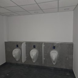 Miller. Bud. Oleh Miller - Trzy białe pisuary zamontowane na szarej, marmurkowej ścianie w publicznej toalecie, z widoczną instalacją i przegrodami oddzielającymi.