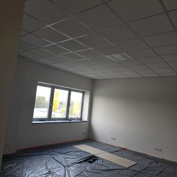 Miller. Bud. Oleh Miller - Pokój w trakcie prac wykończeniowych: zabezpieczona folią podłoga, świeżo pomalowane ściany, sufit z kasetonów, okno z widokiem na drzewa.