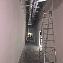 Miller. Bud. Oleh Miller - Korytarz w trakcie prac wykończeniowych, widoczne instalacje pod sufitem, ściany po szpachlowaniu, drabina aluminiowa i sylwetka osoby w oddali.