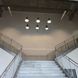 Surowe betonowe schody z metalową balustradą w jasnym wnętrzu, oświetlone wiszącymi lampami i listwami LED na suficie.