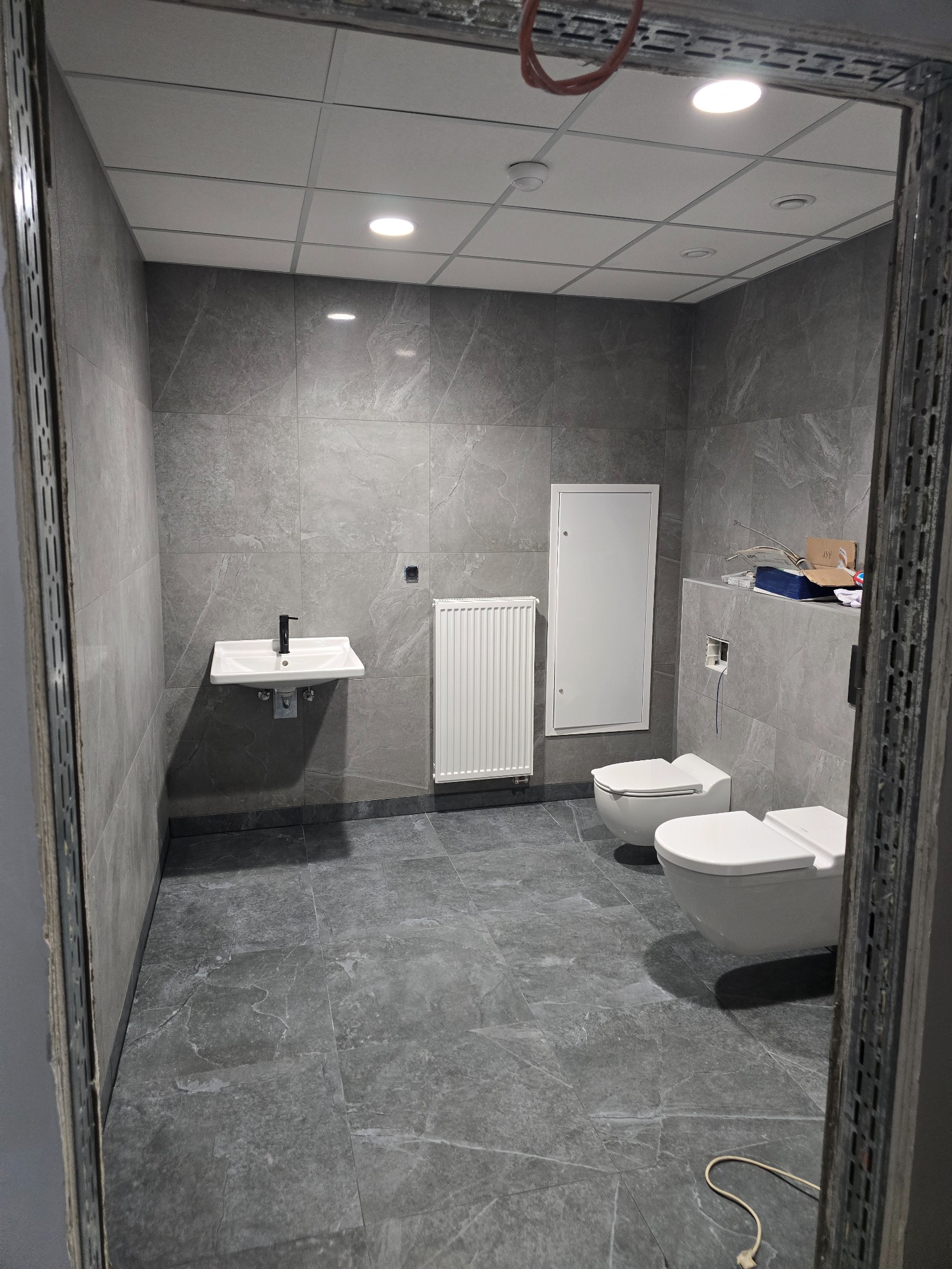 Szara łazienka z dwiema toaletami, umywalką i grzejnikiem, widok przez metalową futrynę w trakcie remontu, widoczne płytki imitujące marmur.