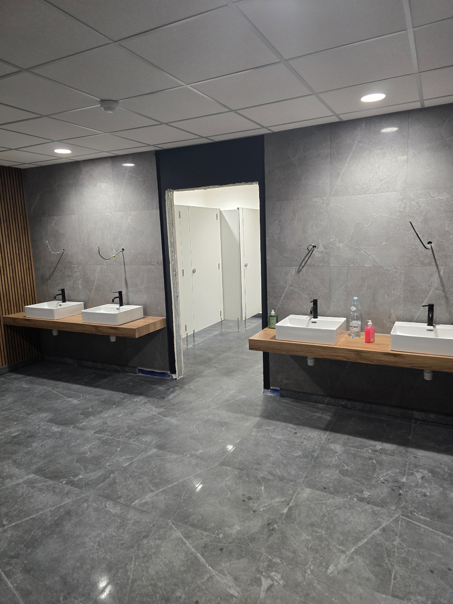 Nowoczesna łazienka publiczna z dwoma umywalkami nablatowymi na drewnianych blatach, szare płytki imitujące marmur na ścianach i podłodze, widoczne instalacje hydrauliczne i wieszaki na ręczniki...