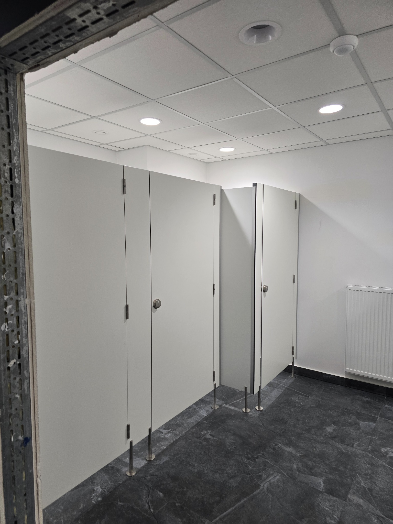Nowe kabiny toaletowe z szarej płyty, zamontowane na metalowych nóżkach, widok z perspektywy wejścia, z widoczną instalacją sufitową i grzejnikiem.