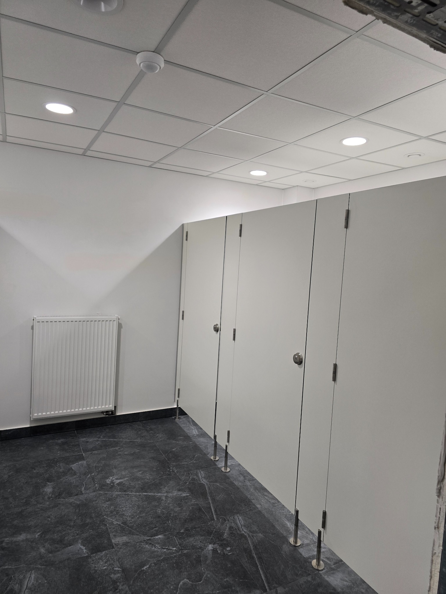 Wnętrze publicznej toalety z kabinami o szarych ścianach, ciemną marmurową podłogą, białym grzejnikiem i sufitem podwieszanym z oświetleniem punktowym.