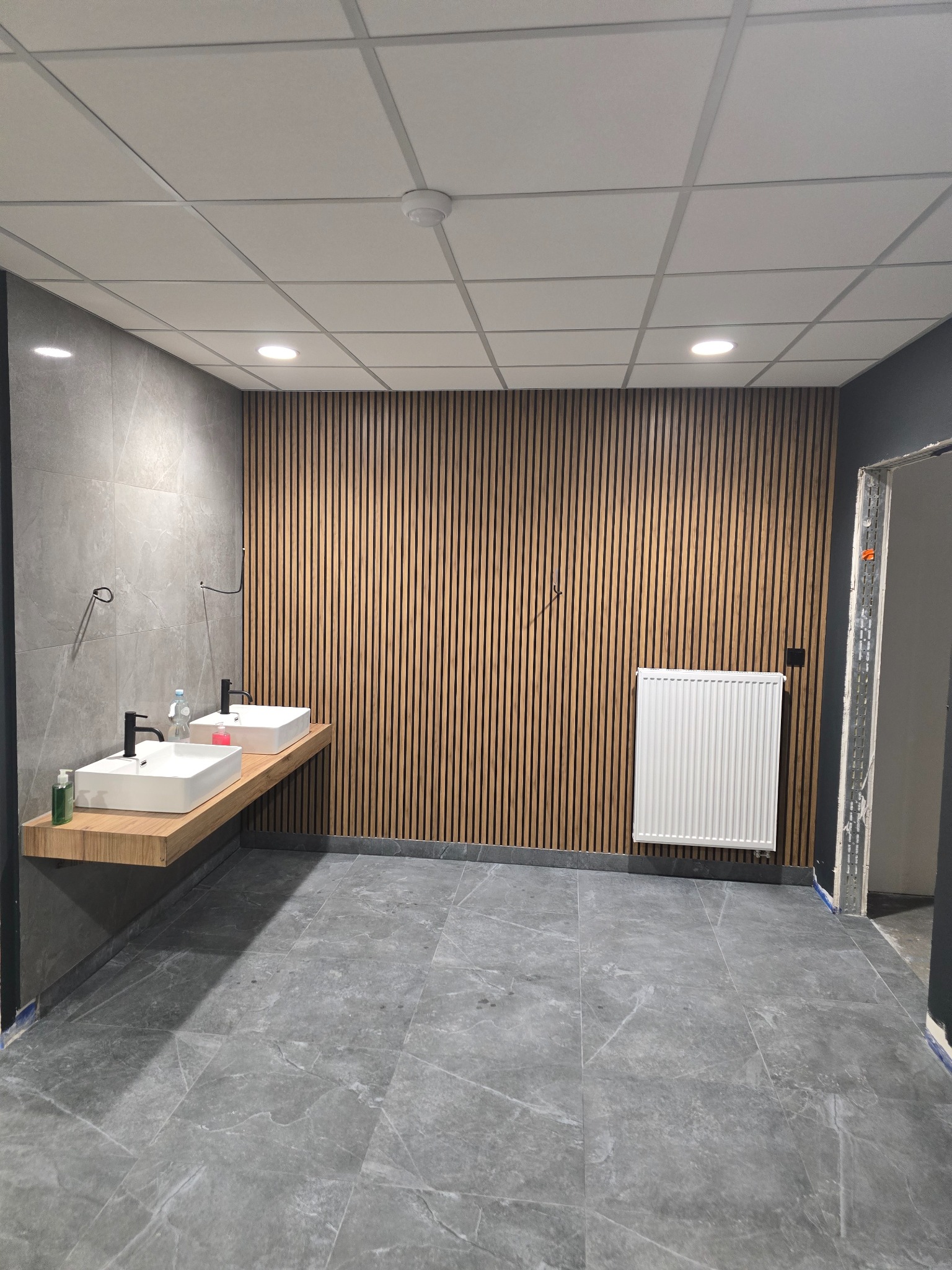 Nowoczesna łazienka z dwiema kwadratowymi umywalkami na drewnianym blacie, drewniana ściana z pionowych listew, szare płytki podłogowe i grzejnik w Piasecznie.