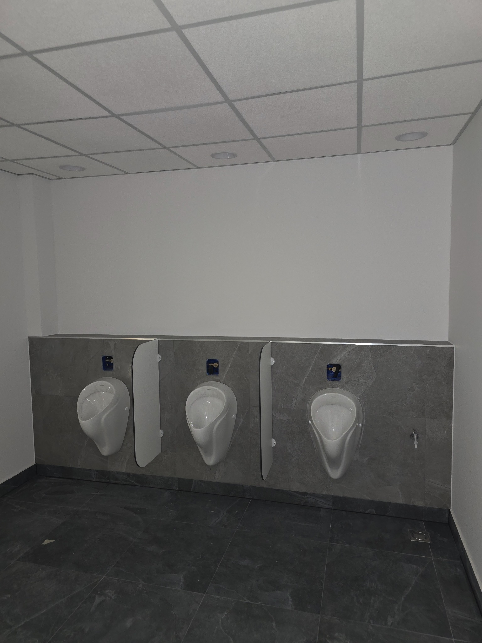 Trzy białe pisuary zamontowane na szarej, marmurkowej ścianie w publicznej toalecie, z widoczną instalacją i przegrodami oddzielającymi.