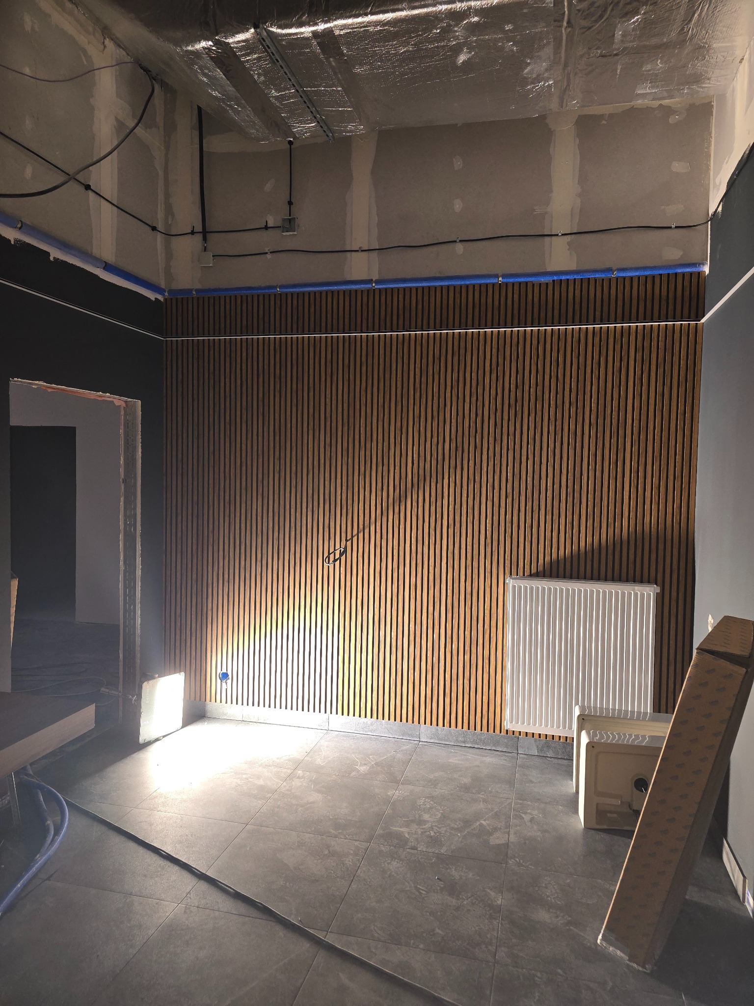 Wnętrze w trakcie prac wykończeniowych: drewniana okładzina ścienna z pionowych listew, grzejnik, surowy sufit z izolacją, podłoga z szarych płytek, instalacje elektryczne i rury wzdłuż ścian.