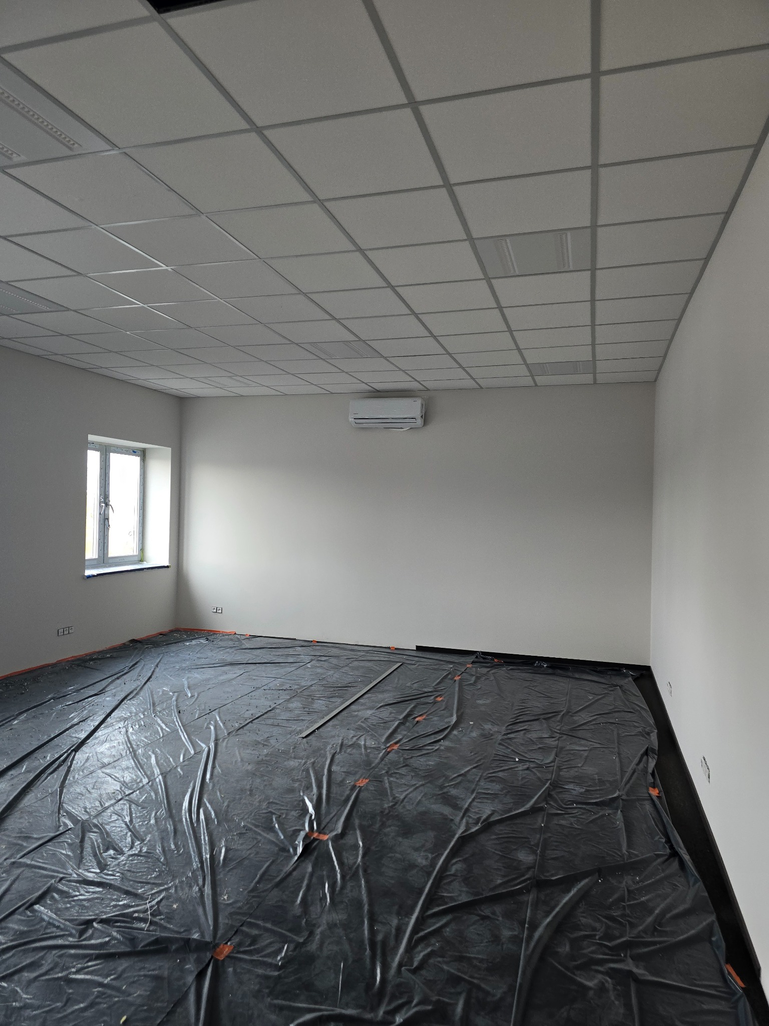 Pokój w trakcie przygotowań do prac wykończeniowych, podłoga zabezpieczona folią, widoczny klimatyzator na ścianie i sufit kasetonowy.