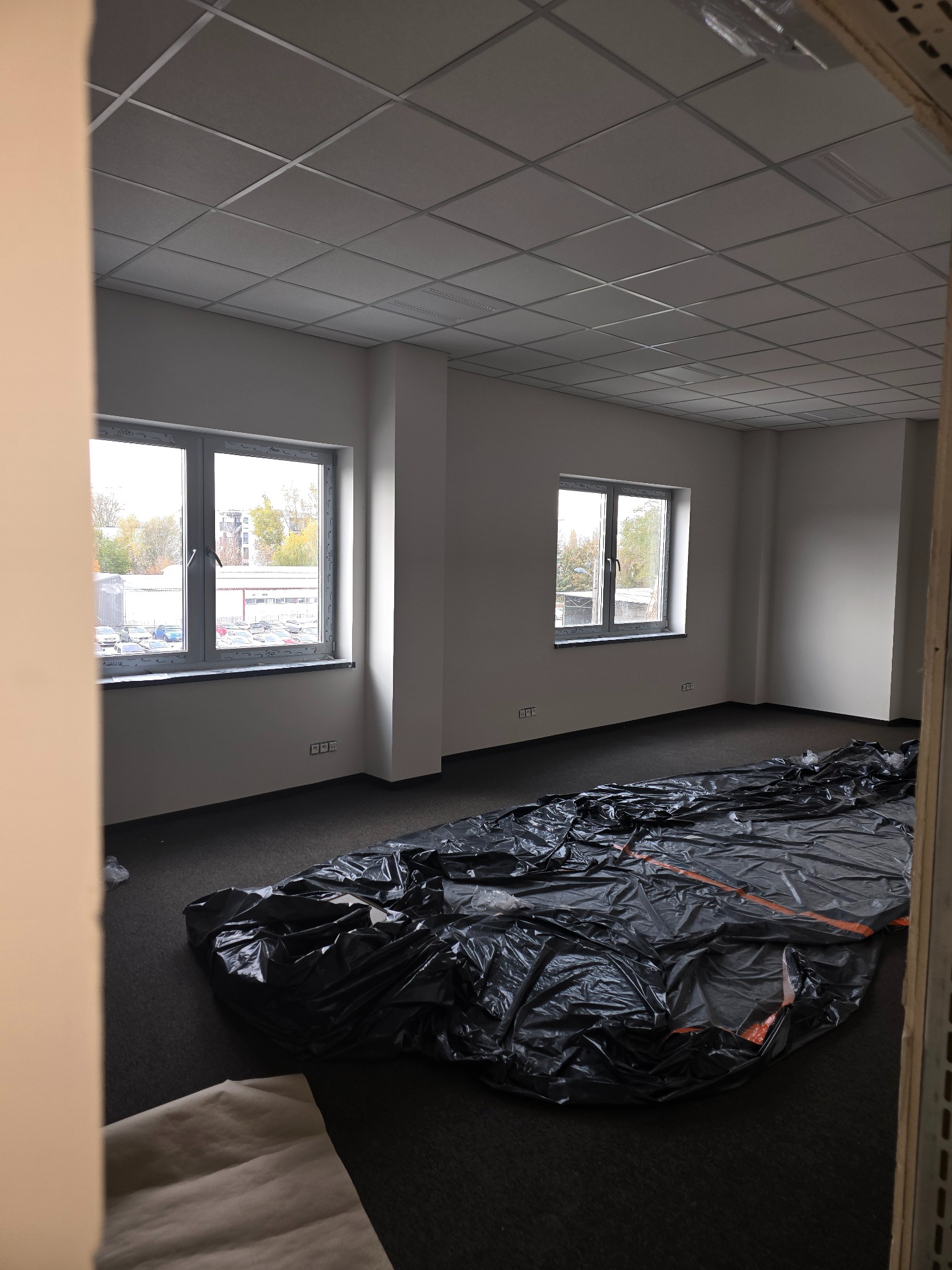 Pusty pokój biurowy z dwoma oknami, zabezpieczony folią malarską przed pracami wykończeniowymi, widok na sufit kasetonowy i fragment ściany w neutralnych barwach.