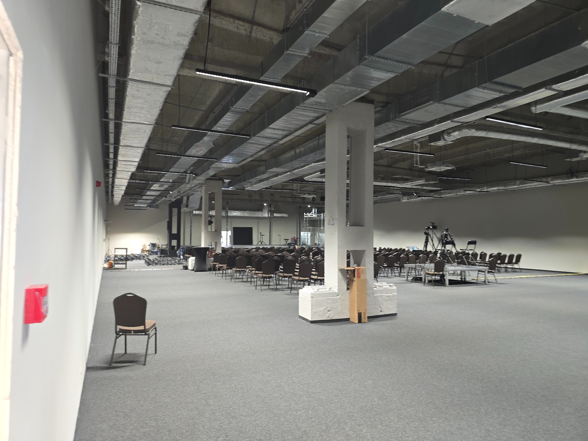 Przestronne wnętrze hali z szarym dywanem, rzędami krzeseł dla publiczności, widocznymi instalacjami pod sufitem i białymi filarami, przygotowane do wydarzenia.
