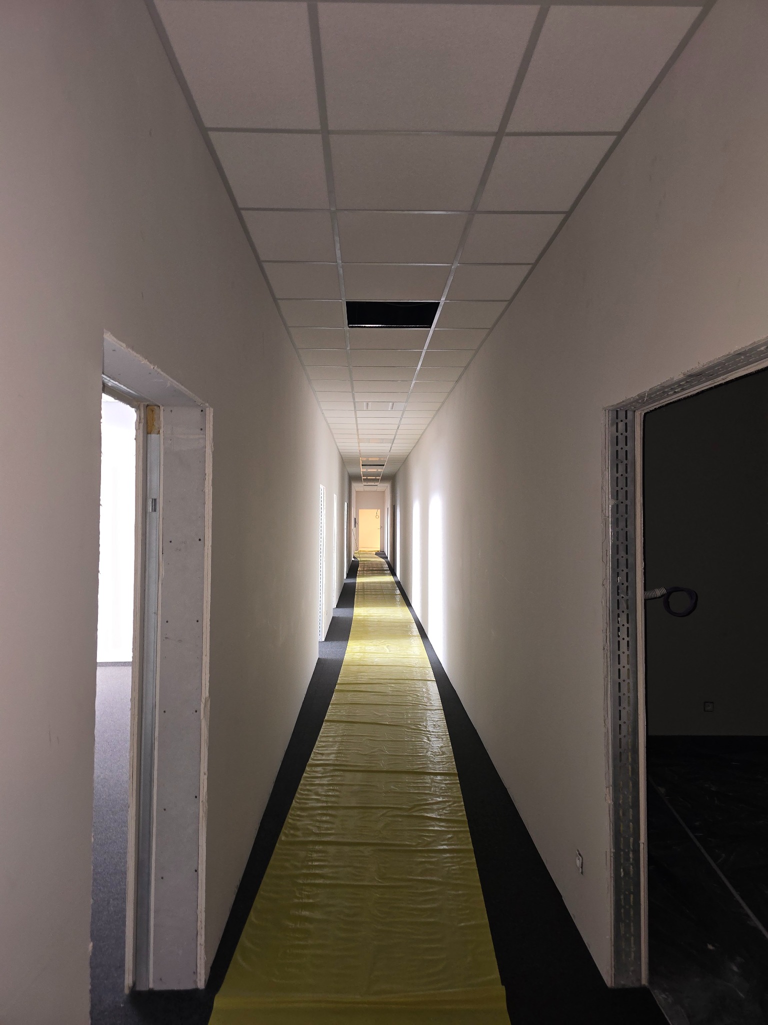 Długi korytarz w biurze w trakcie prac wykończeniowych, z zabezpieczoną folią podłogą, widoczne profile przy ościeżnicach drzwiowych i suficie podwieszanym.