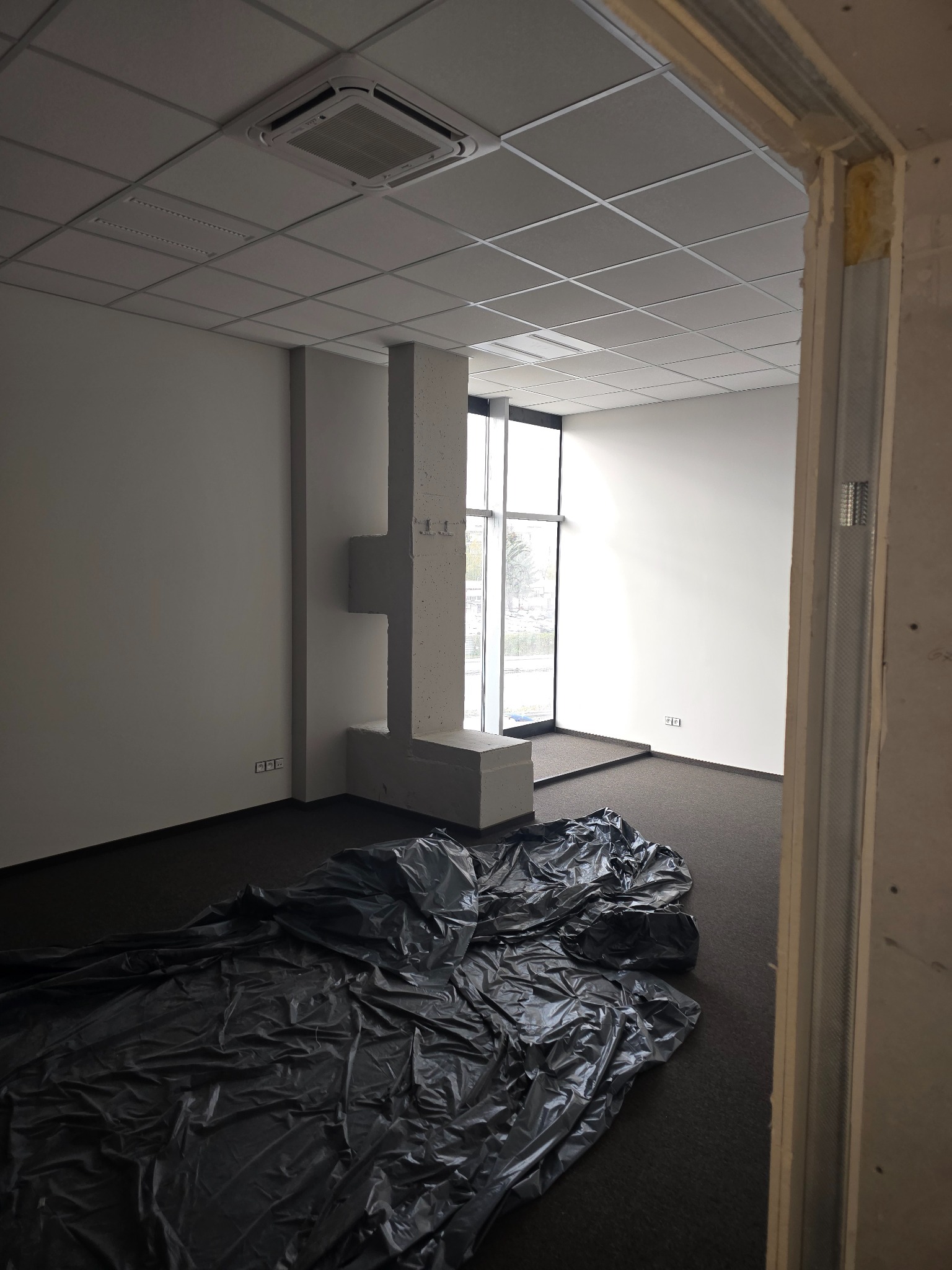 Przygotowanie do prac wykończeniowych w biurze: zabezpieczona podłoga folią malarską, widoczne profile do montażu ścianek działowych, sufit podwieszany i kolumna z elementami dekoracyjnymi.