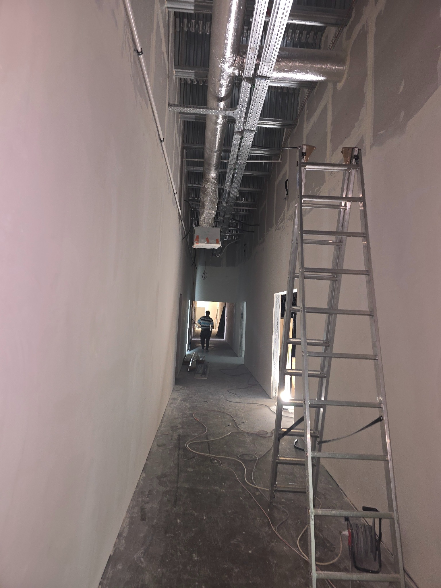 Korytarz w trakcie prac wykończeniowych, widoczne instalacje pod sufitem, ściany po szpachlowaniu, drabina aluminiowa i sylwetka osoby w oddali.