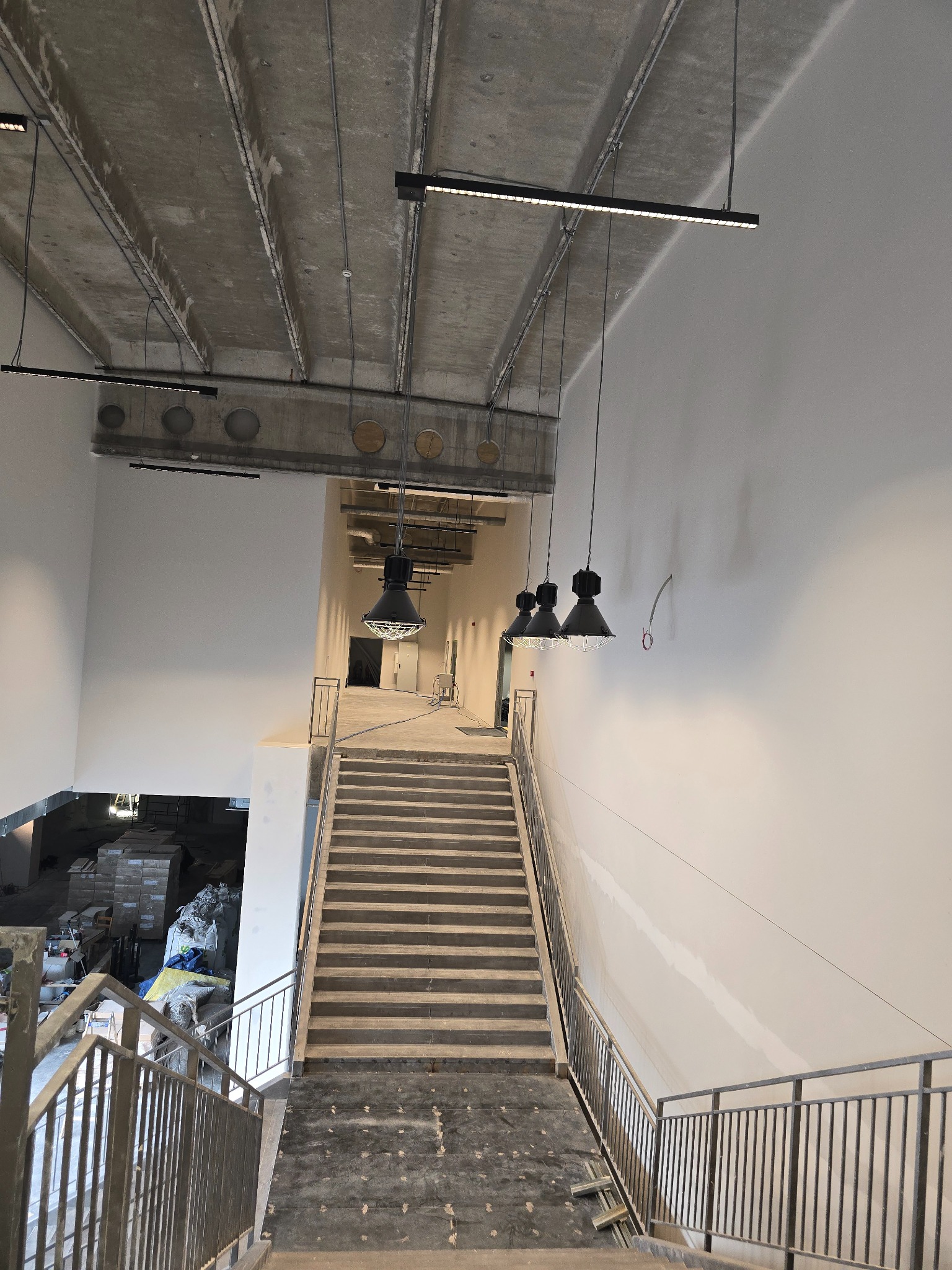 Wnętrze budynku w stanie deweloperskim: surowy betonowy strop z oświetleniem, schody z metalową barierką, ściany przygotowane do malowania, widoczne instalacje i materiały budowlane w tle.