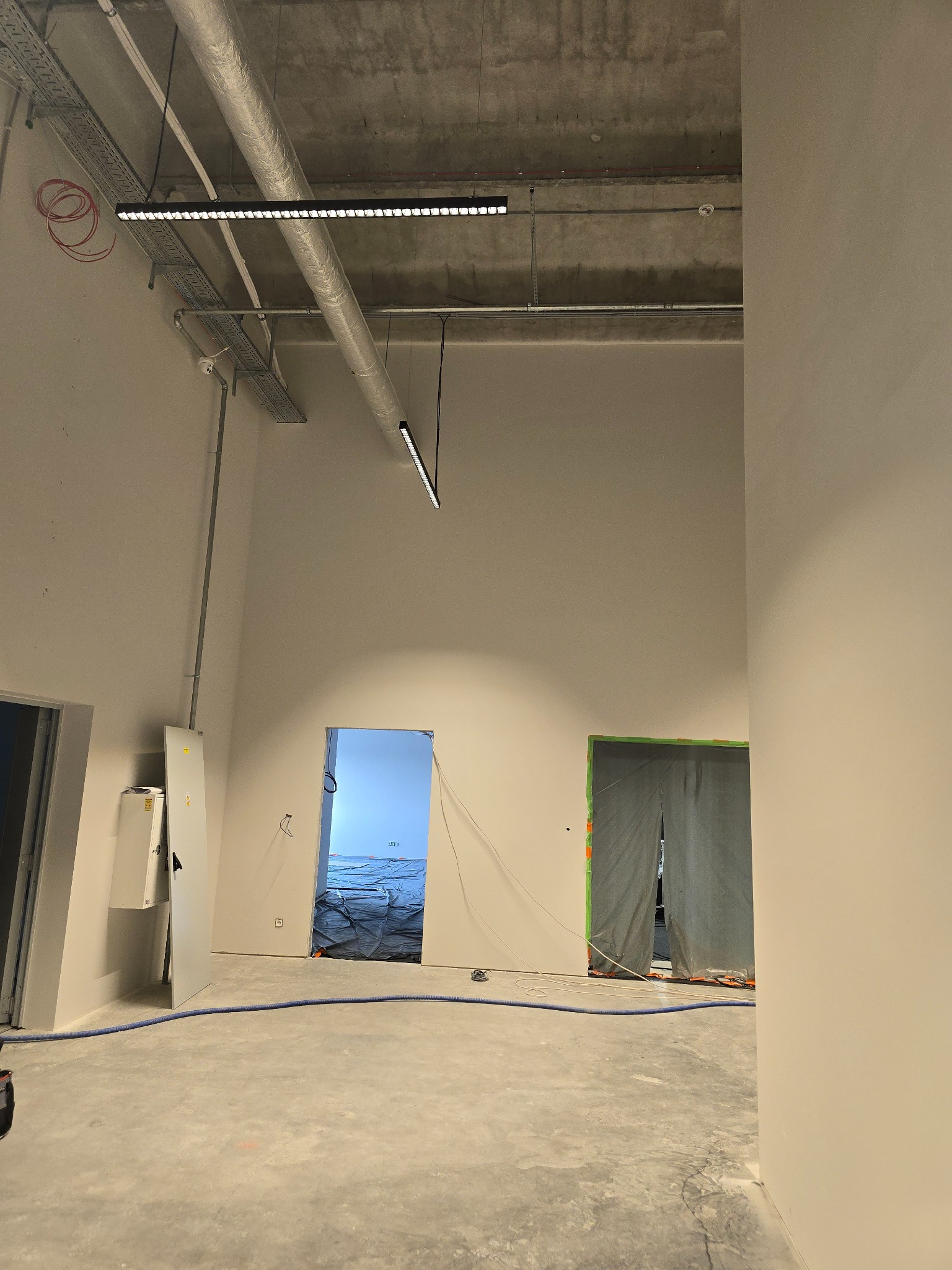 Wnętrze w stanie surowym z widocznymi instalacjami sufitowymi, zabezpieczonymi folią otworami drzwiowymi i stertą materiałów budowlanych w tle, niebieski wąż na podłodze.