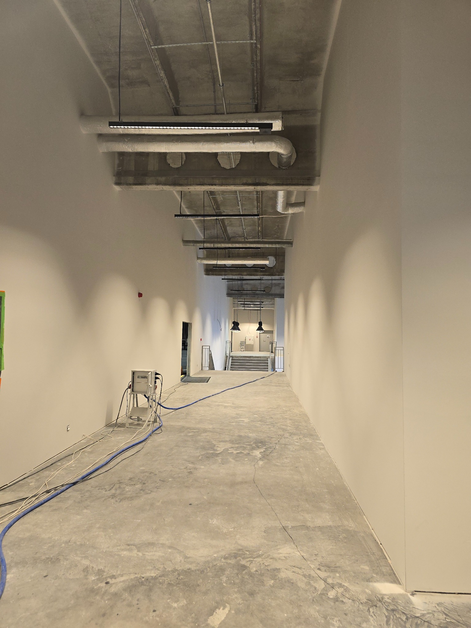Długi korytarz w surowym stanie wykończenia, z widocznymi instalacjami pod sufitem, świeżo pomalowanymi ścianami, betonową posadzką i sprzętem budowlanym pod ścianą.