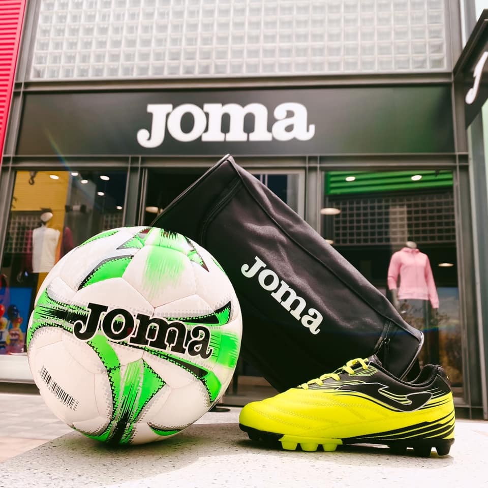 Piłka nożna Joma, żółto-czarne buty piłkarskie i torba sportowa Joma na tle sklepu firmowego Joma z przeszkloną fasadą.