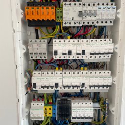 MAREK KALICIAK - Zielona Energia - Rozdzielnica elektryczna z zabezpieczeniami przepięciowymi, bezpiecznikami, okablowaniem i oznaczeniami obwodów fotowoltaicznych, zamontowana w białej obudowie.